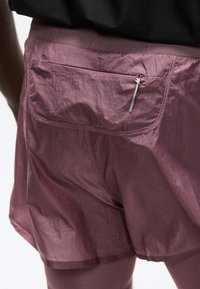 Gros plan sur une jupe de sport bordeaux avec une poche arrière zippée et des coutures décoratives, portée par-dessus un legging assorti.