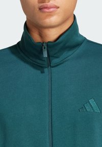 Veste zippée teal avec un col haut, texture lisse et un logo Adidas subtil de couleur assortie sur la poitrine gauche.