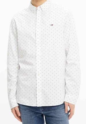 Person iført en hvid skjorte med små mørke firkantede mønstre og blå jeans, hænder afslappet langs siderne.