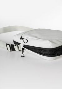 Weiße Umhängetasche mit einem schwarzen Reißverschlussfach, flacher rechteckiger Form, glatter Textur, verstellbarem Schultergurt und Metallakzenten.