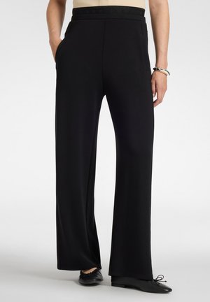 Persoon draagt zwarte wijde broek met elastische tailleband, handen deels in de zakken, gecombineerd met zwarte platte schoenen en zilveren armband.