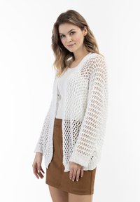 Witte, opengewerkte cardigan met lange mouwen, voorzien van een ontspannen pasvorm en ribgebreide zoom, gecombineerd met een licht tanktop en een bruine rok.