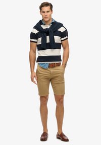 Herenoutfit met een marineblauw en wit gestreepte polo, kaki shorts, een bruine riem en bruine instappers, met een marineblauwe trui over de schouders gedrapeerd.