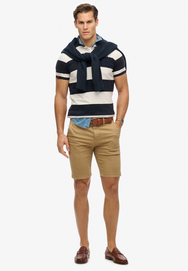 Herenoutfit met een marineblauw en wit gestreepte polo, kaki shorts, een bruine riem en bruine instappers, met een marineblauwe trui over de schouders gedrapeerd.