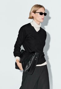 Maglione nero a scollo a v con un dolcevita bianco, abbinato a pantaloni neri a gamba larga. Il modello porta una grande borsa in pelle nera.