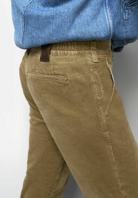 Beige Cordhosen mit einer leicht strukturierten Oberfläche, elastischem Bund und einem braunen Logo-Patch auf der Rückseite. Sichtbare Seitentaschen.
