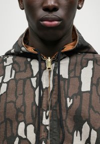 Hoodie in Braun und Creme mit einem abstrakten Muster, ausgestattet mit einem goldenen Reißverschluss und schwarzen Kordeln. Das Material wirkt strukturiert.