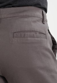 Pantalon en coton gris présentant un design épuré, une texture subtile, et une poche arrière avec une bordure cousue. Finition du tissu lisse.