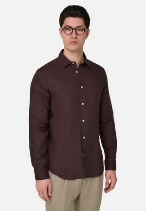 Jeune homme portant une chemise marron foncé boutonnée et un pantalon beige, debout devant un fond clair uni, faisant face à l'avant.
