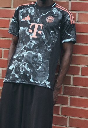 Personne portant un maillot de sport noir et gris à motifs du FC Bayern Munich avec des logos et des rayures corail, debout devant un mur en briques rouges.