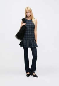 Marineblaues ärmelloses Kleid mit Karomuster, ausgestelltem Saum und Schnallenakzenten; kombiniert mit dunkelblauen ausgestellten Jeanshosen und einer schwarzen Kunstpelzhandtasche.