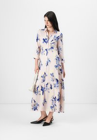Knälång klänning med knappar i lätt rosa med blå blommönster. Har trekvartsärmar och midja som är cinchad, kombinerad med svarta slingback-skor.