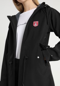 Giacca nera con cappuccio, chiusura a zip, vita regolabile con cordino e tasche laterali. Presenta un design con una patch rossa sul petto e una texture morbida.