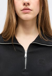 Νεαρή γυναίκα που φοράει μαύρο επάνω μέρος με φερμουάρ σε ασημένιο χρώμα, με εμφανή τα χείλη, το πιγούνι και μέρος από τα μακριά καστανά μαλλιά της, πάνω σε λευκό φόντο.