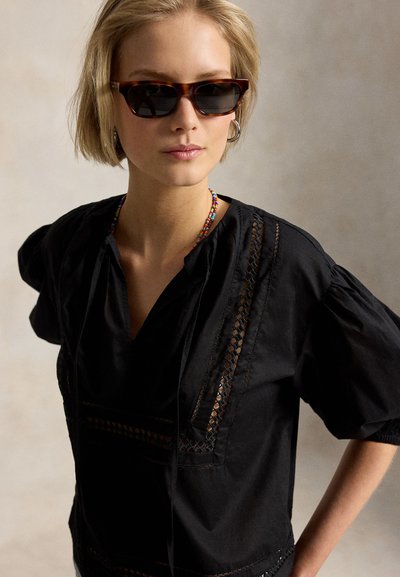 Ξανθιά γυναίκα με σκούρα γυαλιά ηλίου με μοτίβο χελώνας, μαύρο μπλουζάκι με λεπτομέρειες από δαντέλα, πολύχρωμο κολιέ με χάντρες και κρίκους σκουλαρίκια σε ουδέτερο φόντο.