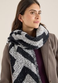 Svart och vit scarf med zigzag-mönster, tillverkad i ett strukturerat stickat material, med fransar i kanten och som hänger löst runt halsen.