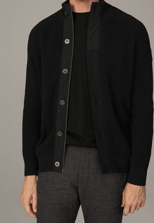 Homme portant un cardigan noir côtelé à boutons par-dessus une chemise noire et un pantalon à carreaux gris, debout devant un fond gris uni.