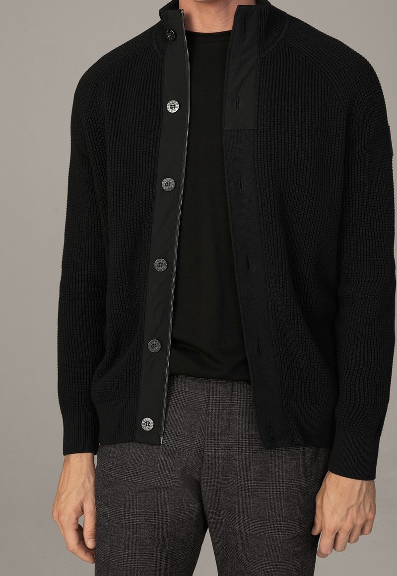 Homme portant un cardigan noir côtelé à boutons par-dessus une chemise noire et un pantalon à carreaux gris, debout devant un fond gris uni.