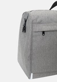 Grå tygväska med en strukturerad yta, vinklad form, svart hårdvara och en liten vit detalj på nederkanten.