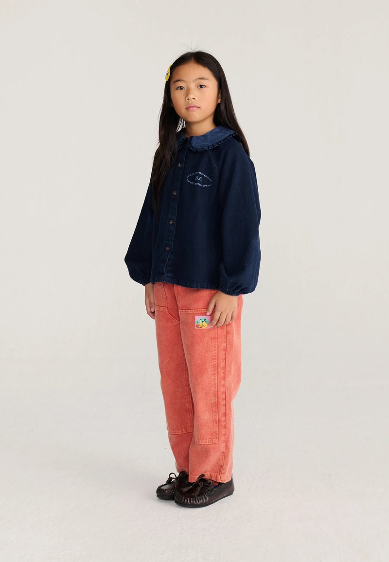 Jeune fille aux longs cheveux noirs portant une chemise bleu marine boutonnée et un pantalon corail ample avec un patch coloré, debout sur un fond clair.