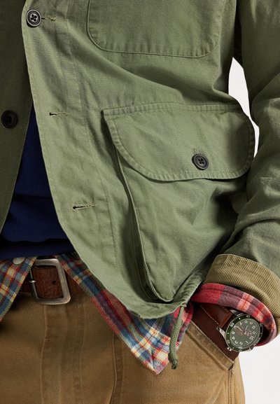 Μπουφάν ελιάς με τσέπη στο στήθος, συνδυασμένο με πουκάμισο φανέλα καρό σε κόκκινο, μπλε και πράσινο. Διαθέτει καφέ ζώνη και μαύρο ρολόι στον καρπό.