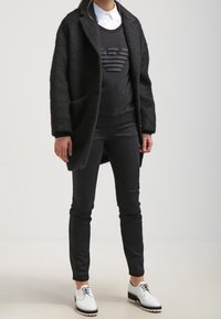 Svart, fuzzy oversized kappa över en svart tröja med en texturerad design, åtsittande svarta byxor och vita skor med svarta detaljer.