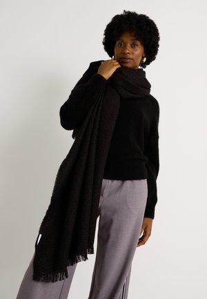 Bufanda negra de punto con flecos, llevada sobre un suéter negro y combinada con pantalones gris claro, que presentan una textura sutil.