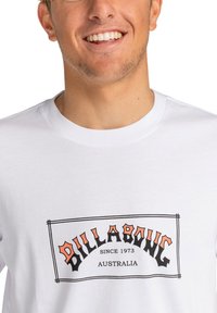 Bílý bavlněný tričko s obdélníkovou grafikou, na které je černý a oranžový text s nápisem "BILLABONG SINCE 1973 AUSTRALIA" v retro stylu.