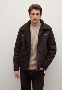 Braune Shearling-Jacke mit einem strukturierten Kragen und dezenten Lederakzenten. Getragen über einem hellbeigen, grob gestrickten Pullover und dunkelbraunen Hosen.