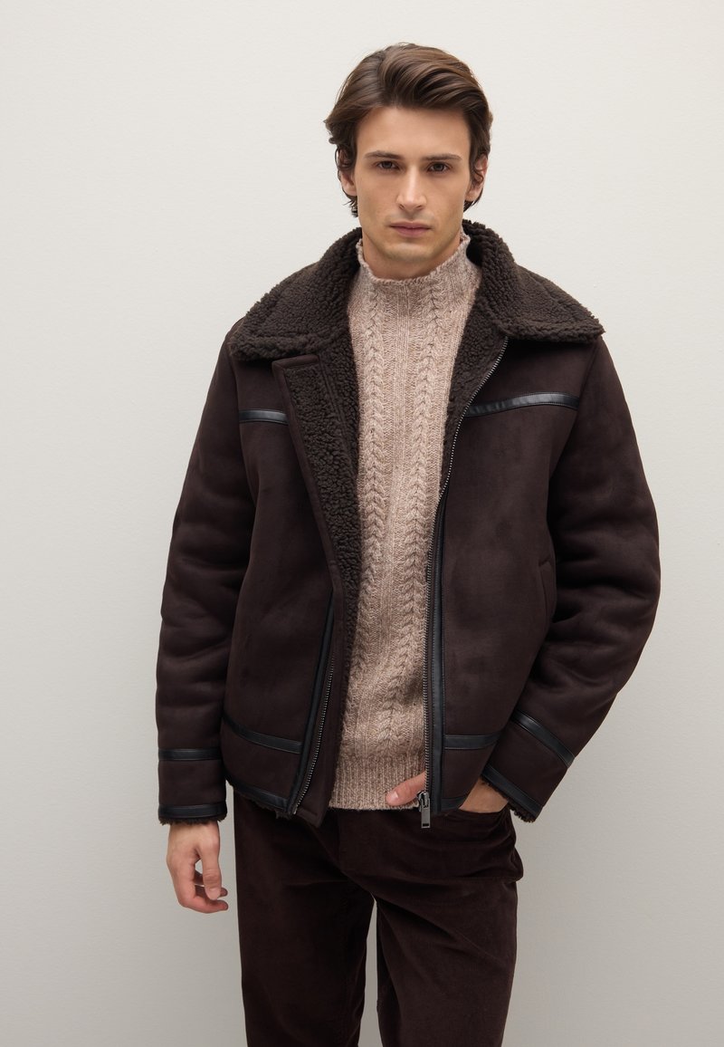 Braune Shearling-Jacke mit einem strukturierten Kragen und dezenten Lederakzenten. Getragen über einem hellbeigen, grob gestrickten Pullover und dunkelbraunen Hosen.