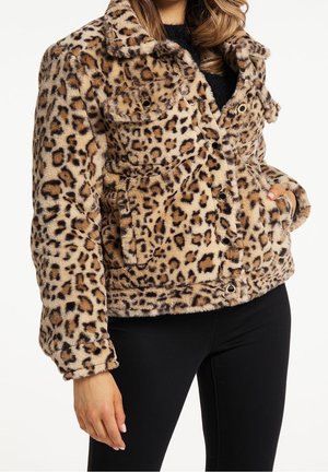 Jakke med leopardprint i falsk pels med trykknapper, båret over sort top og sorte bukser, modelens hånd i lommen og løst bølget hår synligt.