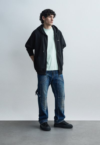 Νεαρός άνδρας με σγουρά σκουρόχρωμα μαλλιά, φοράει μαύρο φούτερ με φερμουάρ, ανοιχτόχρωμο πουκάμισο, φαρδιά μπλε cargo τζιν και μαύρα αθλητικά παπούτσια, στέκεται σε γκρι πάτωμα.