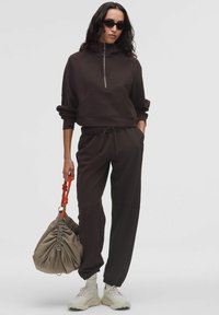 Nő sötétbarna zipzáras kapucnis pulóverben és melegítőnadrágban, fehér sportcipőben, fekete napszemüvegben, egy bézs zsinóros táskával, amelynek narancssárga fogói vannak.