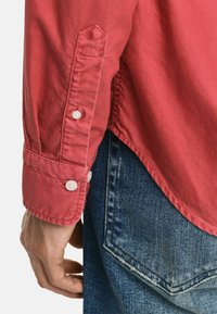 Rotes Hemd mit Knopfleiste und umgeschlagenen Ärmeln, ausgestattet mit weißen Knöpfen und einer weichen Textur, kombiniert mit blauen Jeans.