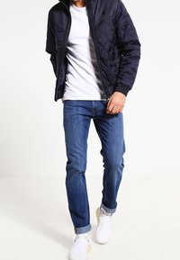 Marinblå quiltad jacka med hög krage, vit t-shirt och blå jeans. Modell bär ljusa sneakers och står på en neutral bakgrund.