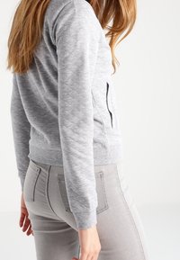 Kvinna i en tight, ljusgrå, vadderad jacka och ljusgrå jeans som står i profil med hand avslappnat vid sidan.