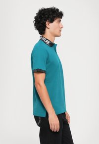 Ung mann i teal kortermet polo med svart krage og ermekanter, stående i profil mot en enkel hvit bakgrunn.