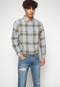 Camisa de franela a cuadros en colores azul, verde y amarillo; con dos bolsillos frontales; combinada con jeans azul desgastados. Ajuste general relajado.