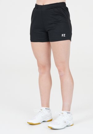 Person iført sorte træningsshorts med "FZ"-logo og hvide sportssko med gule såler, stående mod en ensfarvet baggrund.