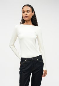 Λευκή ριμπ μπλούζα μακρύ μανίκι με φαρδύ ντεκολτέ, που διαθέτει μικρό λογότυπο στο αριστερό στήθος, σε συνδυασμό με σκουρόχρωμο τζιν παντελόνι.