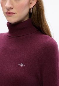 Dunkelburgunderfarbener Turtleneck-Pullover mit geripptem Halsausschnitt und gesticktem Logo auf der Brust. Weicher, texturierter Stoff.