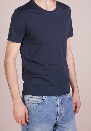 Mann trägt ein schlichtes dunkelblaues T-Shirt mit kurzen Ärmeln und hellblaue Jeans, steht vor einem neutralen Hintergrund.