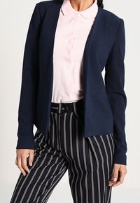 Blazer bleu marine avec une coupe ajustée, manches longues, ouverture sur le devant, associé à un polo rose clair et un pantalon à rayures noires et blanches.