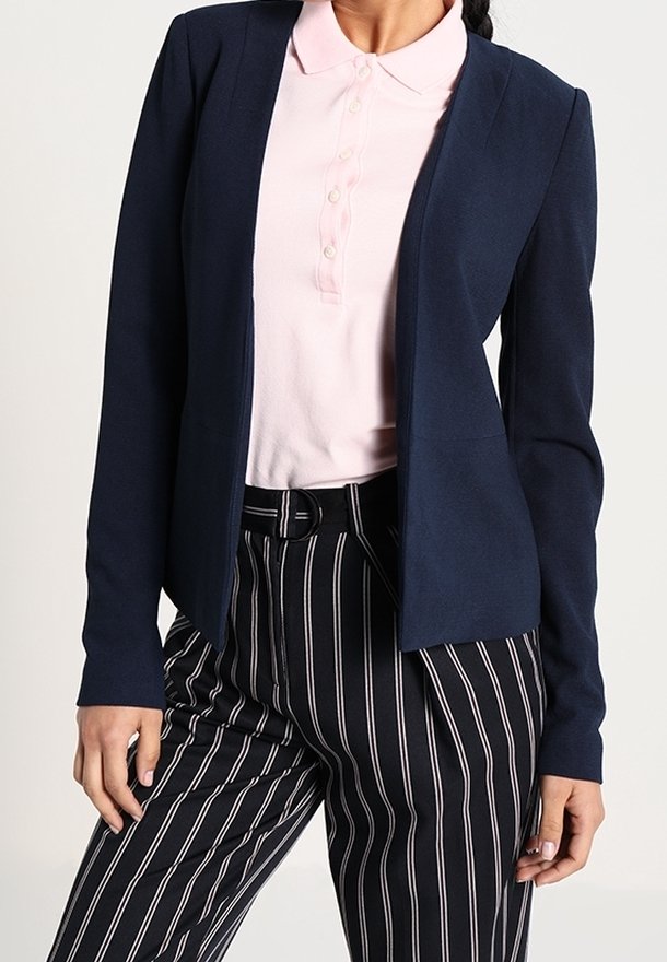 Blazer bleu marine avec une coupe ajustée, manches longues, ouverture sur le devant, associé à un polo rose clair et un pantalon à rayures noires et blanches.