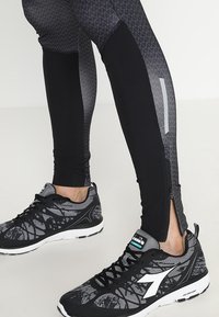 Jambes portant des leggings noirs et gris à motifs avec zips au niveau des chevilles, associés à des chaussures de sport noires et grises avec semelles blanches et logos.