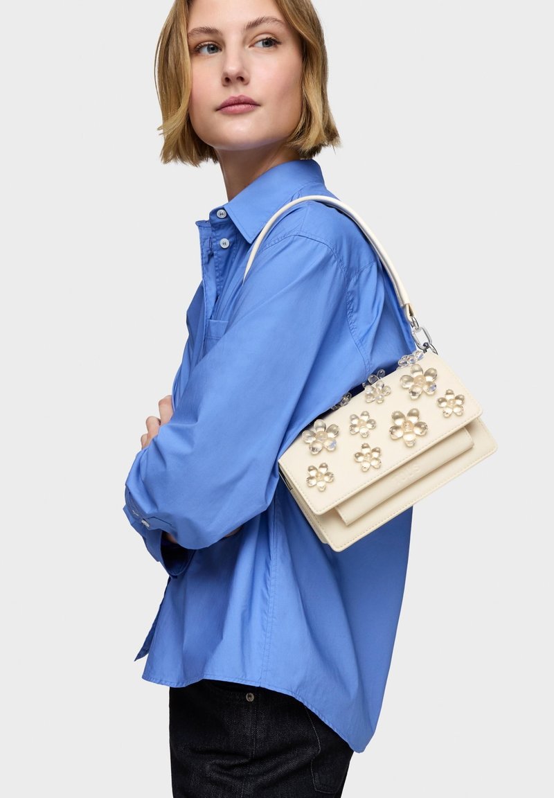 Sac à main beige avec ornements floraux en perles, design structuré et bandoulière amovible, tenu contre une chemise bleue.
