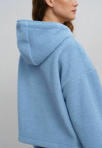 Lys blå fleece hættebluse med en tekstureret overflade, oversized pasform og foret hætte, der viser et minimalistisk design uden synligt hardware.