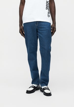 Mees, kes kannab sirgeid siniseid teksapükse, valget t-särki musta tekstiga, valgeid sokke ja must-valgeid loafereid, seisab ühevärvilise tausta ees.