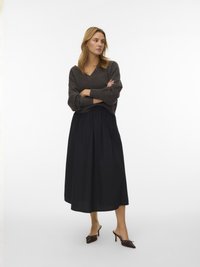 Pull en maille gris foncé à col en V, associé à une jupe midi plissée noire taille haute. Escarpins marron à bouts pointus avec des accents décoratifs.