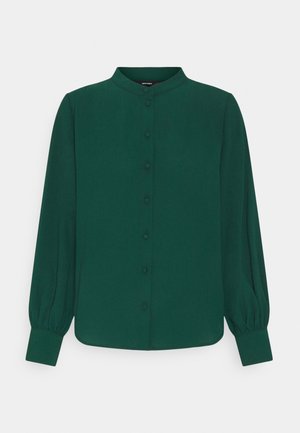 Mørkegrøn bluse med lange ærmer, rund krave, knapper foran og rynkede manchetter på en ensfarvet lys baggrund.